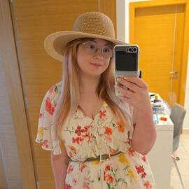 Leah is taking a mirror selfie, she us wearing a large sunhat and a white floral dress.